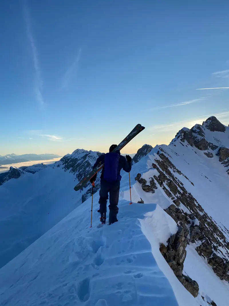 Un skieur monte à pied sur une arête étroite et enneigée, skis sur l'épaule