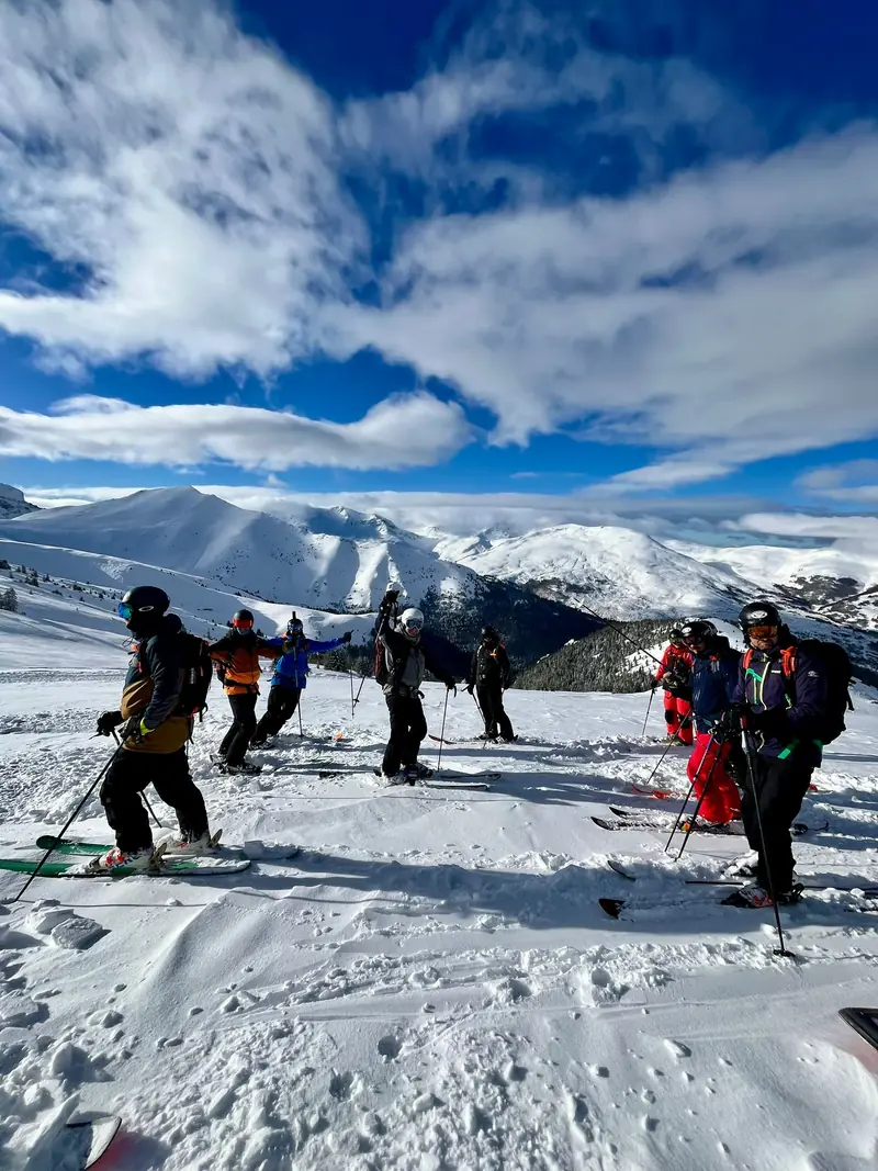 A group of skiers and snowboarders gathered on a summit plateau under dramatic clouds
