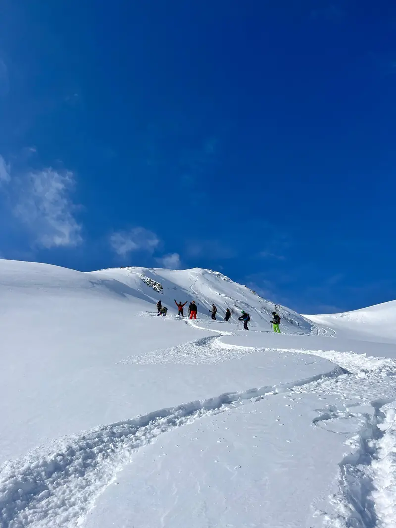 A line of ski tourers climbing a wide snowy slope under a blue sky