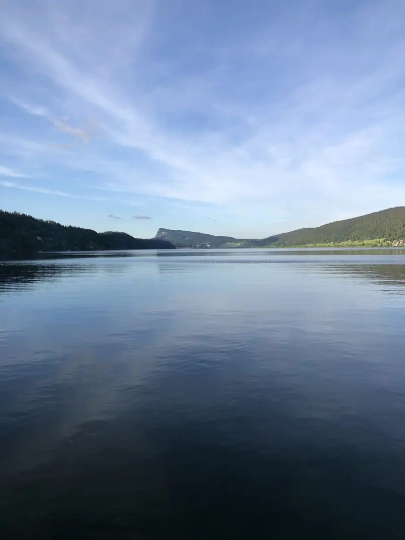 Ein stiller See, in dem sich bewaldete Hügel und der Sommerhimmel spiegeln